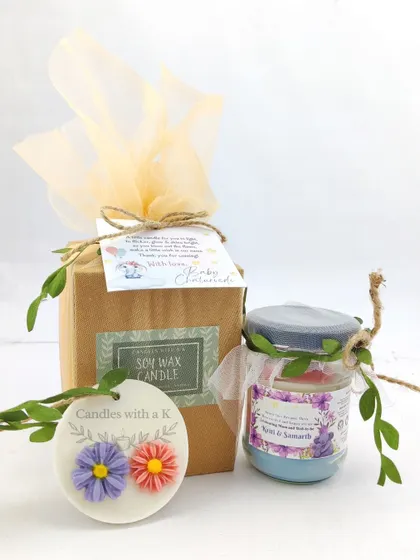 A baby shower return gift box containing a layered 100ml soy wax candle and a mini floral wax sachet.