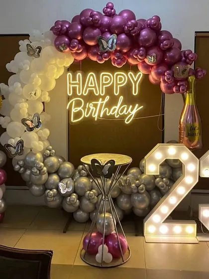 A chic 23rd birthday decoration with a semi-arch of purple, silver, and white balloons. The setup is completed with a neon sign, marquee numbers, and champagne-themed foil balloons.