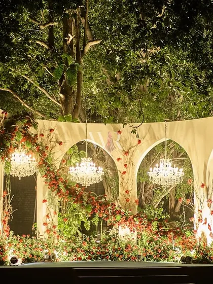 This romantic stage, set against a backdrop of old trees, was designed for an engagement. We used white arches draped with cascading red and green florals, with chandeliers nestled within to create a magical, garden-like setting.