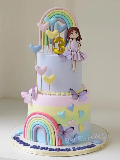 A sweet and colorful two-tier cake featuring rainbows, butterflies, and a cute fondant girl topper. The pastel palette and heart accents make this a lovely choice for a young child's birthday.