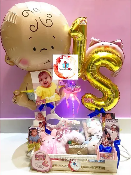 A grand first birthday hamper for a baby girl. It's decorated with large foil balloons, personalized photos, and filled with toys and accessories.