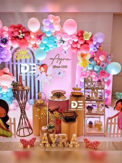 A vibrant fairy-themed first birthday for Ayra. This colorful setup includes large paper flowers, a rainbow balloon arch, and cute fairy characters, creating a joyful and playful atmosphere.