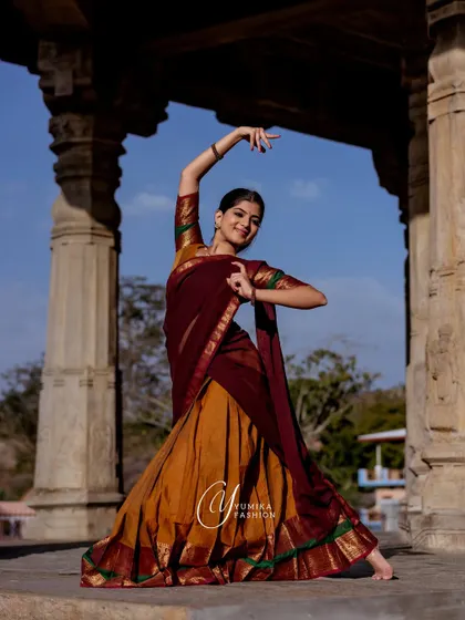 A powerful dance pose in a mustard and maroon half-saree, framed by the grand pillars of a temple. This image demonstrates the dramatic potential of combining dance and fashion.