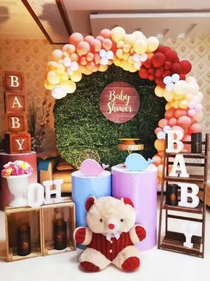 A modern and cute baby shower decoration. This setup uses a green foliage backdrop, a balloon arch in warm tones, and adorable props like a teddy bear and 'Oh Baby' signs.