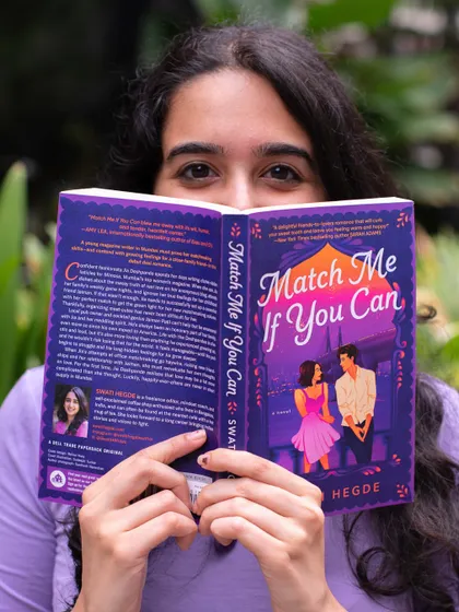 Peeking over a copy of my debut novel, *Match Me If You Can*. This book is a desi romance pitched as *Emma* meets *When Harry Met Sally*.