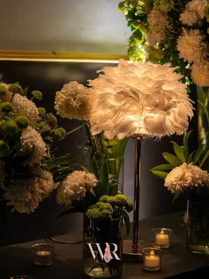 Ambiance is everything. A feather-adorned lamp casts a soft, diffused light over arrangements of white chrysanthemums, creating a moody and textural detail in a lounge corner.