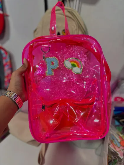This transparent pink backpack is customized with a 'P' initial and a cute rainbow patch. It's a fun and trendy accessory for kids and adults alike.