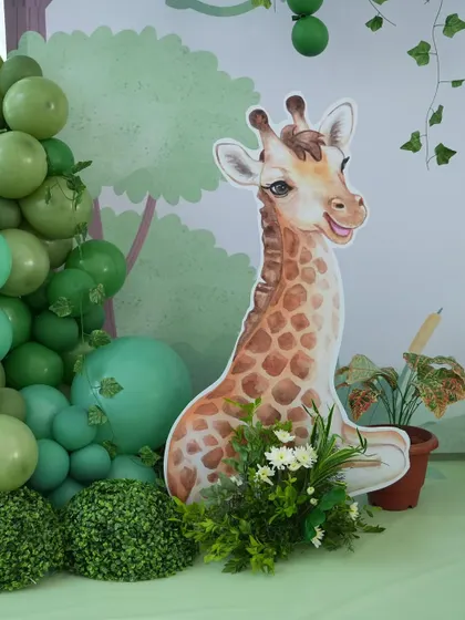A friendly giraffe cutout peeks out from behind a lush arrangement of greenery and a balloon garland, adding a touch of playful charm to this wild-themed birthday party.