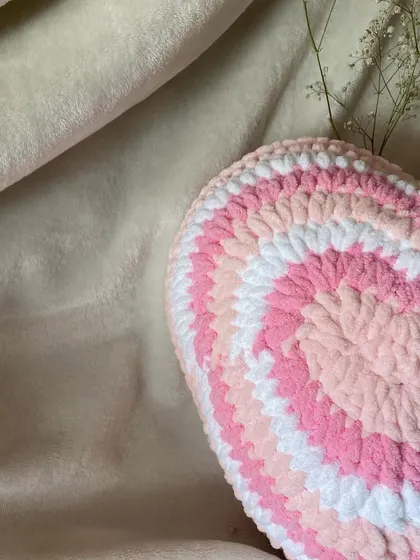 A styled shot of our pink and white crochet heart pillow. The soft, plush texture makes it a perfect decorative accent for a cozy corner of your room.