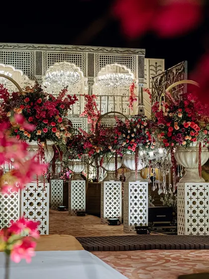 A closer view of the floral arrangements lining the aisle, with the intricate jaali-work backdrop visible behind. The vibrant pinks and reds of the flowers pop against the white pedestals.