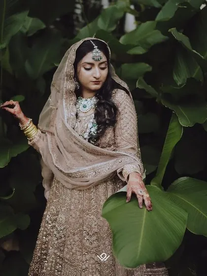 A Muslim bride in a stunning beige lehenga, captured in a natural outdoor setting. The contrast between her attire and the lush green leaves creates a striking image.