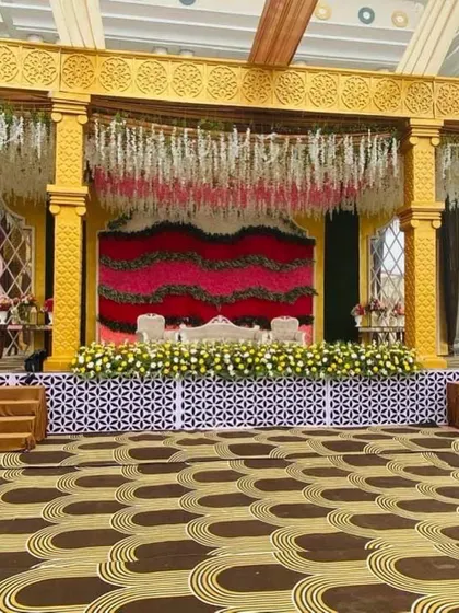A full view of a reception stage with a unique patterned floor. The backdrop features waves of red and pink flowers, complemented by hanging floral arrangements and traditional pillars.