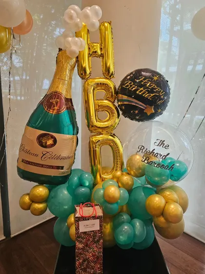 A celebratory balloon bouquet perfect for an adult's birthday. This arrangement features a large champagne bottle foil balloon, 'HBD' letters, and a mix of teal and gold balloons.