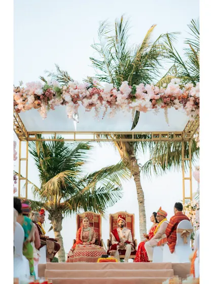 The couple seated at their mandap during a picturesque beachside ceremony in Dubai, framed by palm trees and a floral arch.