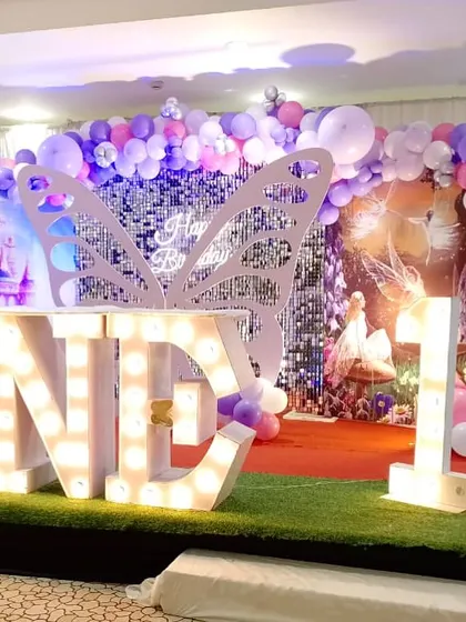 A whimsical first birthday party with a fairy theme. The stage features a large silver butterfly prop, "ONE" marquee letters, and a fairy cutout, creating an enchanting atmosphere.