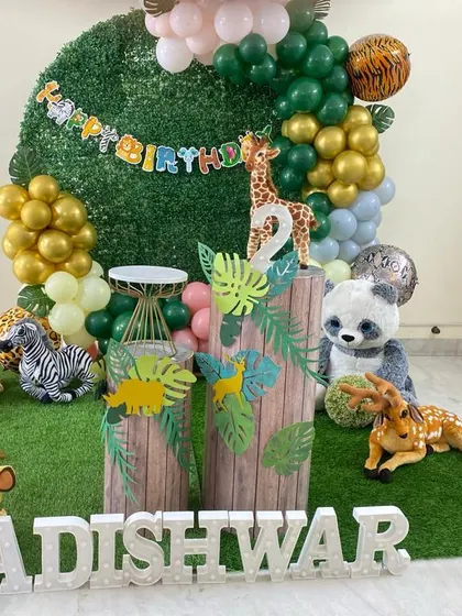 A close-up of a jungle-themed setup on a faux grass mat. The decor includes stuffed animals, a green and gold balloon arch, and wood-textured plinths.