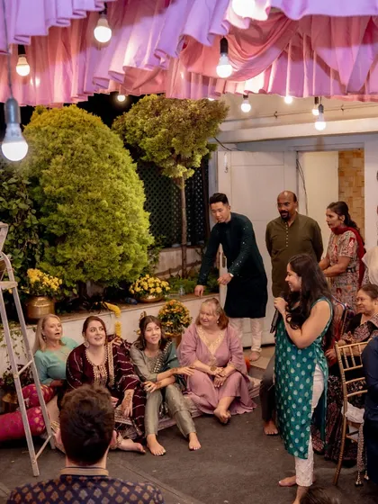 An intimate family gathering during the Mehendi celebrations, with guests seated on the floor, sharing stories and laughter under a canopy of pink drapes and lights.