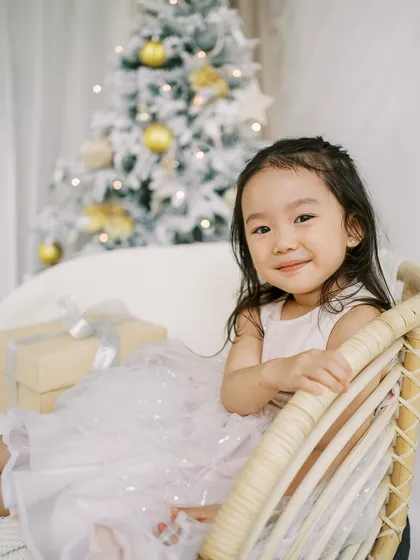 A little girl in a beautiful dress, sitting in a wicker chair by the Christmas tree. A classic and elegant holiday portrait.