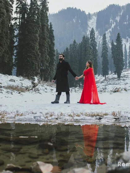 A beautiful reflection shot of the couple holding hands by a calm lake in Kashmir. The snow-covered trees and mountains are perfectly mirrored in the water, creating a symmetrical and serene image.