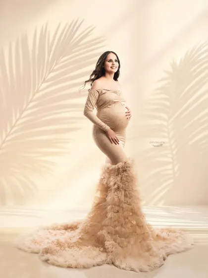 A full-length view of a stunning beige gown with a long, feathery train. The soft, warm lighting and shadow overlays give this studio portrait a gentle and dreamy quality.