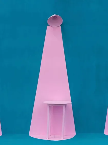 A playful and minimalist installation featuring spotlights and tables in pastel pink against a bold teal wall.