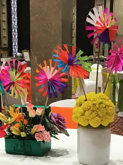 A fun and quirky centerpiece for a Mehendi, featuring colorful paper windmills, marigolds in a ceramic pot, and a floral arrangement in a rustic green trunk.