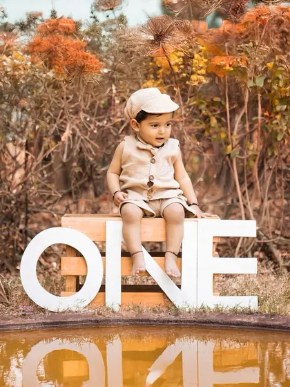An outdoor first birthday shoot with a rustic, adventurous theme. The little one is dressed as an explorer, sitting by a small pond with a "ONE" sign and a tent.