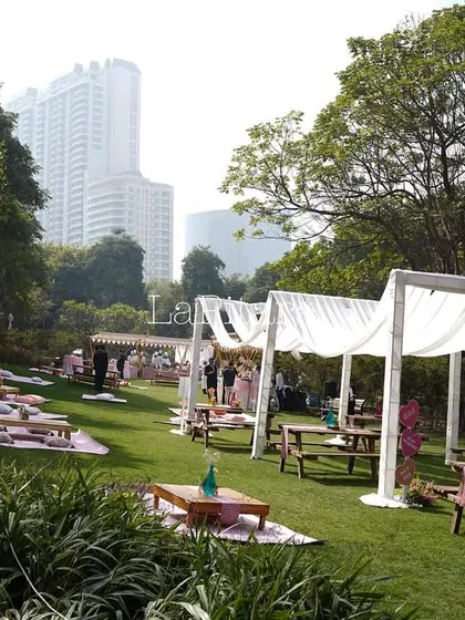 A wide shot of a romantic Valentine's Day picnic setup in a park. The scene features low picnic tables, canopies with white drapes, and plenty of cozy cushions on the grass.