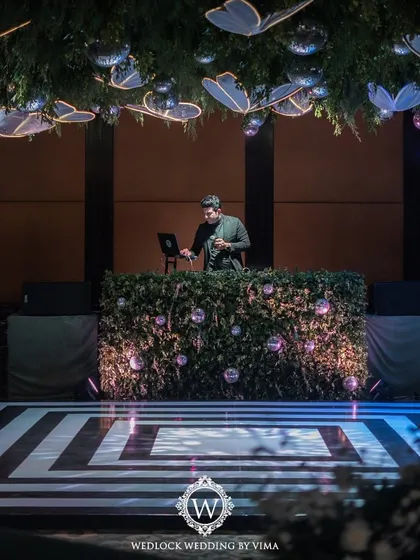 The DJ booth and dance floor for a glamorous Sangeet night. The booth is covered in lush greenery, and the ceiling is adorned with disco balls and glowing butterflies, ready for a night of celebration.