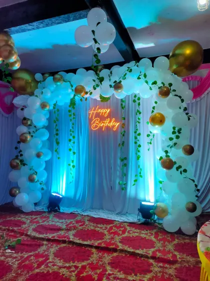 A beautiful backdrop created with white fabric drapes, blue uplighting, and a warm neon 'Happy Birthday' sign, framed by a white and gold balloon arch with greenery.