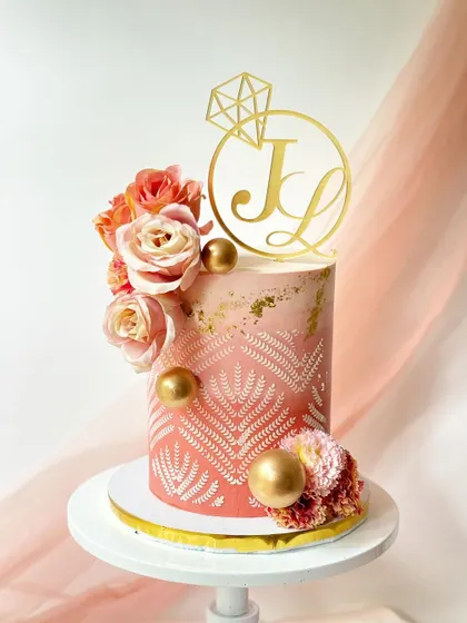 An intricate engagement cake with a modern henna-inspired stencil design. The pink ombre cake is decorated with silk flowers, gold spheres, and a custom initial topper.