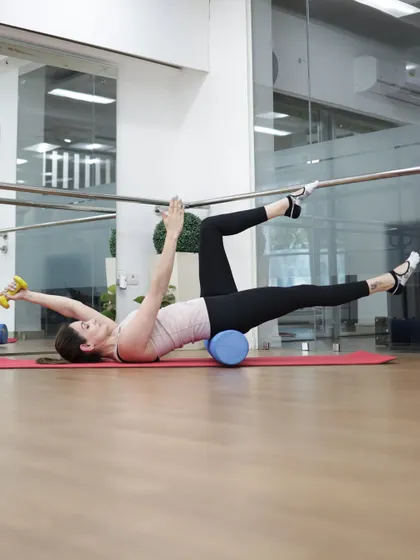 An instructor performs a weighted dead bug on a foam roller, an advanced variation that challenges stability and deep core control.