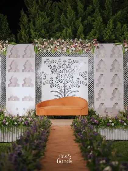 A Sheesh Mahal-inspired reception stage, featuring intricate white panels, a plush orange sofa, and a pathway lined with lavender flowers.