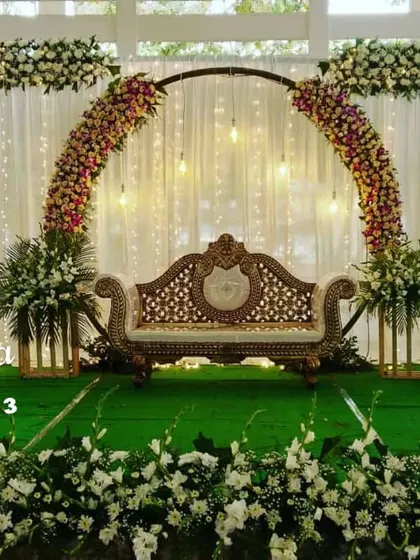 An elegant outdoor stage setup with a lush green turf floor. The design features a semi-circular floral arch and clean white drapery, perfect for a day or evening garden wedding reception.