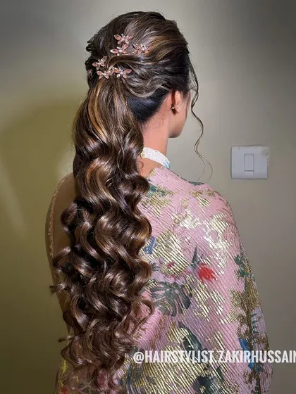 A voluminous ponytail with soft curls and a twisted crown, accented with delicate floral clips. This is a beautiful and elegant style for a reception or engagement party.