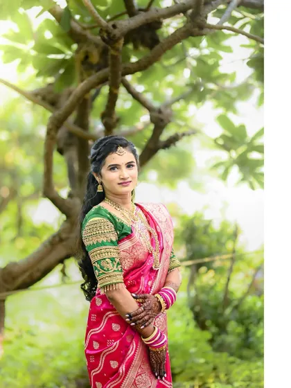 A full-length portrait of the bride in her traditional saree, standing gracefully under a tree. The natural setting highlights the vibrant colors of her outfit.