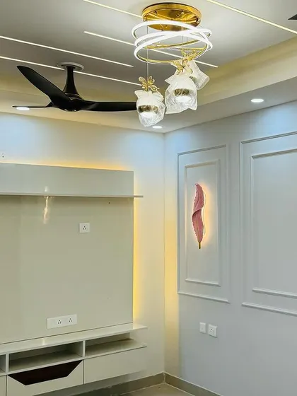 This living room combines a sleek TV unit with a feature wall using POP moulding. The modern chandelier and false ceiling lights complete the look.