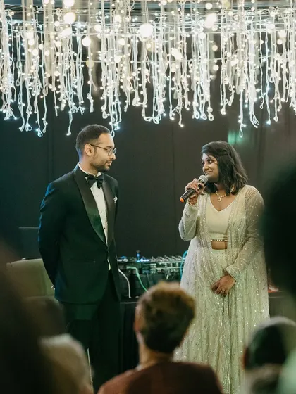 The bride gives a speech at her reception, standing next to her husband under a ceiling of twinkling lights.