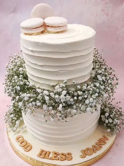 We offer a blessing for a sacred milestone with this elegant two-tiered Holy Communion cake. A wreath of delicate baby's breath separates the textured white tiers, which are topped with pristine white macarons, creating a look of pure serenity.
