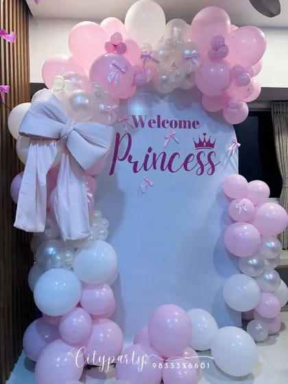 An elegant 'Welcome Princess' decoration for a baby girl's arrival. This home setup features a white arched backdrop, a pink and white balloon arch, and a large fabric bow for a delicate touch.