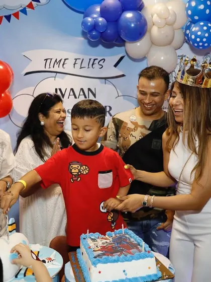 The birthday boy, wearing a crown, cuts his cake at his airplane-themed party, surrounded by his loving family.
