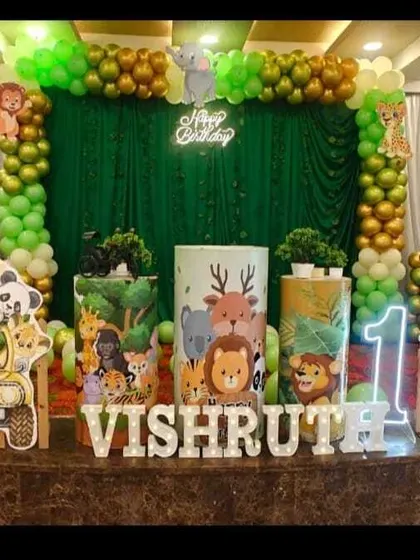A safari-themed first birthday stage. The backdrop features a jeep with cartoon animals, framed by a green balloon arch and marquee number '1'.