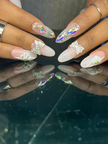 An ethereal and intricate design featuring 3D sculpted flowers, rhinestone bows, and iridescent diamond-shaped gems on long almond nails.