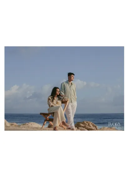 A serene portrait of the couple by the sea in Bali. The composition is clean and minimalist, focusing on their calm presence against the backdrop of the blue sky and ocean, conveying a sense of peaceful togetherness.