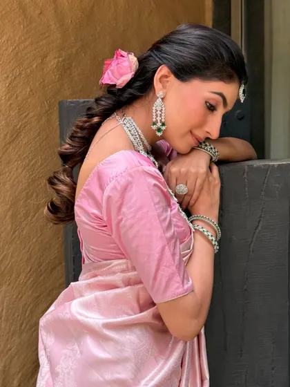 A beautiful side profile showing the romantic braided hairstyle adorned with a fresh pink rose. The makeup is soft and glowing, with a focus on flawless skin.