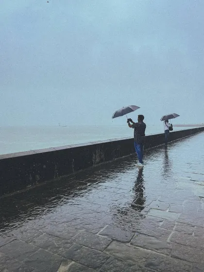Two people brave the wind and rain at the promenade, their umbrellas held against the strong sea breeze. A testament to the spirit of Mumbai.