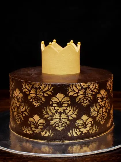 A royal chocolate truffle cake for a special occasion. This egg-free cake features a rich chocolate ganache with a gold damask stencil pattern and is topped with a golden crown. It's filled with fresh strawberries inside.