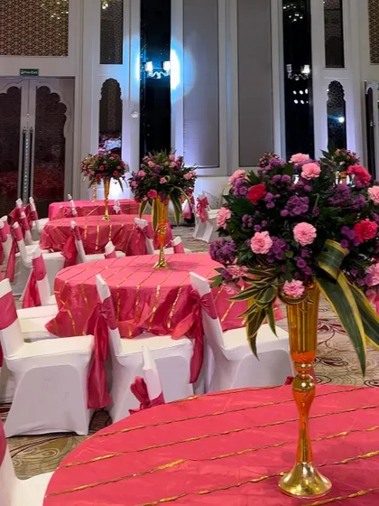 Bright and cheerful table settings for a Mehendi function. The tables are dressed in vibrant pink linens with gold details, topped with tall floral centerpieces in matching hues.