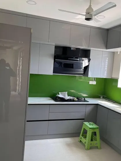 This L-shaped kitchen features glossy grey cabinets and a vibrant green lacquered glass backsplash that adds a pop of color. The tall unit seamlessly integrates the refrigerator.