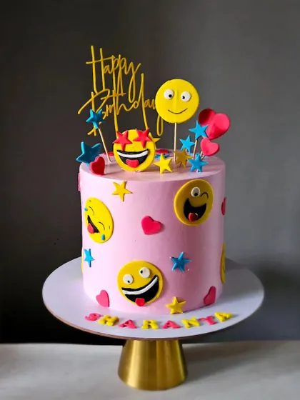 A fun and expressive emoji-themed cake. The pink buttercream is covered in various handmade fondant smiley faces, making it a cheerful and modern cake for a birthday.